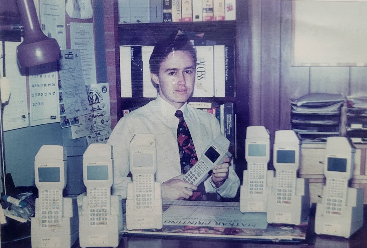 Andrei Spiegel in 1995 in the home office his father built in half the family garage, behind seven Symbol
                  PDT3100 terminals on a desk, holding the seventh