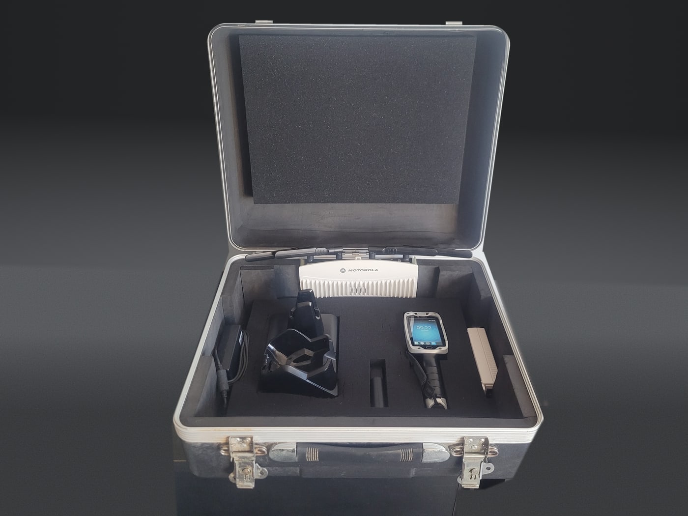 An aluminium flight case open, foam cut to hold a Zebra TC8000 gun-grip
                    terminal, its single-slot charging cradle, a power supply and a spare
                    battery, with the spare access point and its aerials seated across the
                    top of the foam.