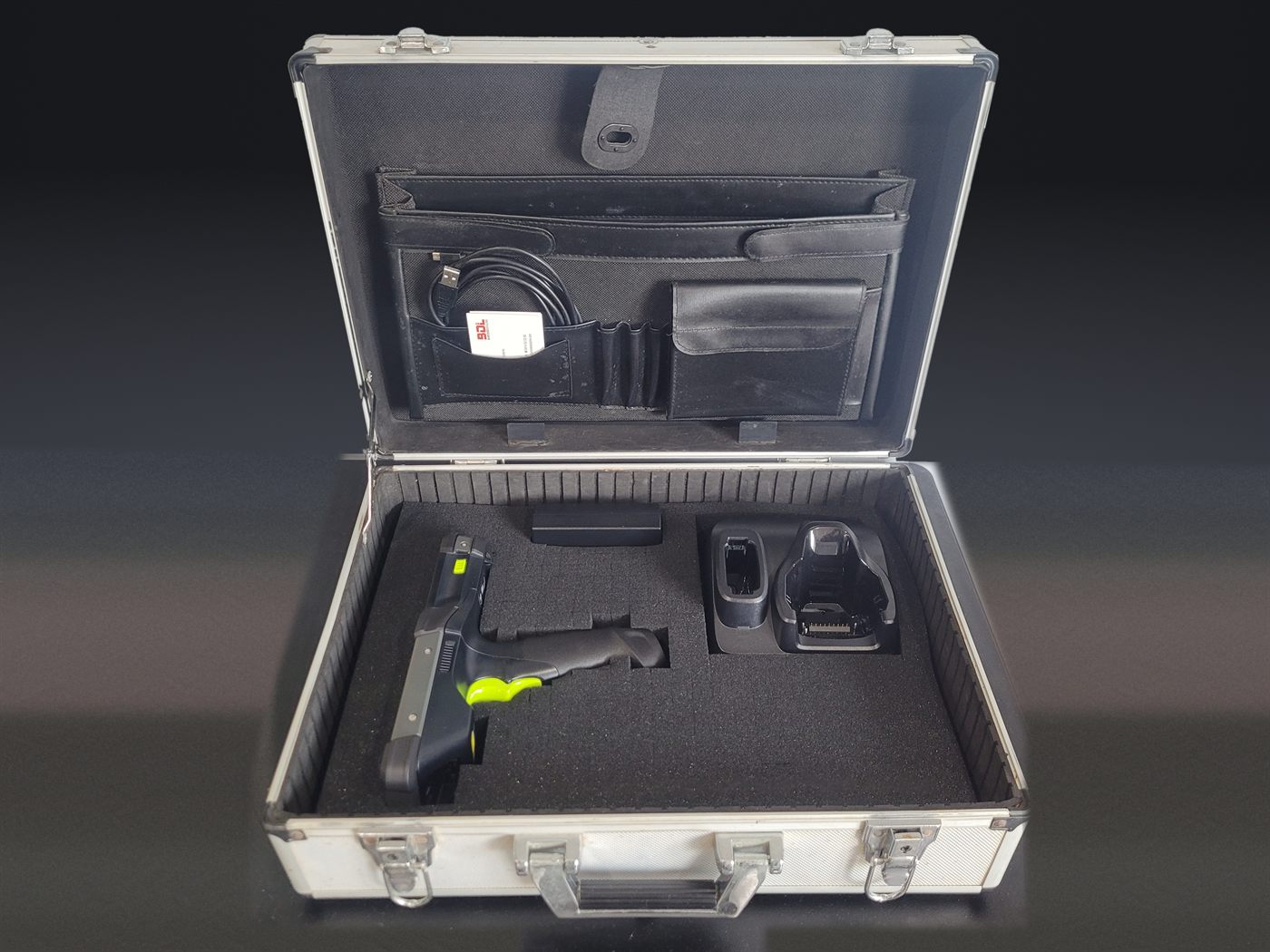 An aluminium flight case open on a bench, foam cut to hold a Zebra MC9400
                    gun-grip terminal, its power supply and a charging cradle, with a USB cable
                    partly visible in the lid pocket and a Barcode Datalink card beside it.