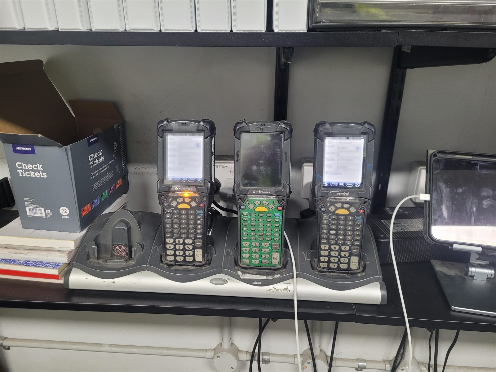Three Motorola MC9100-series handheld terminals standing in a Symbol four-bay charging cradle on a shelf, all visibly worn from years of use. Their screens are pixelated.