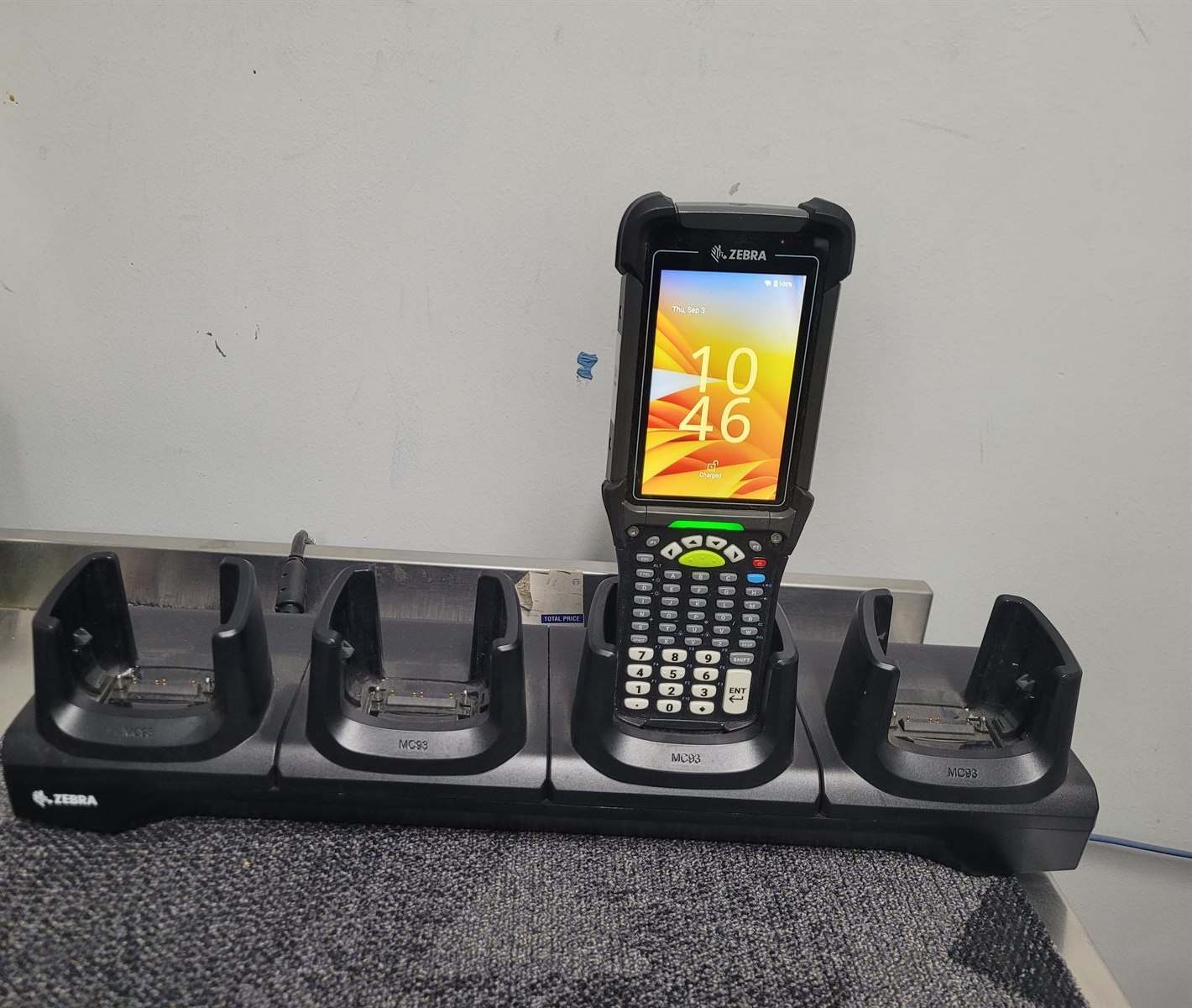 A Zebra MC9400 sitting charged in a black four-slot Zebra cradle moulded MC93, on a stainless steel bench in a food processing area.