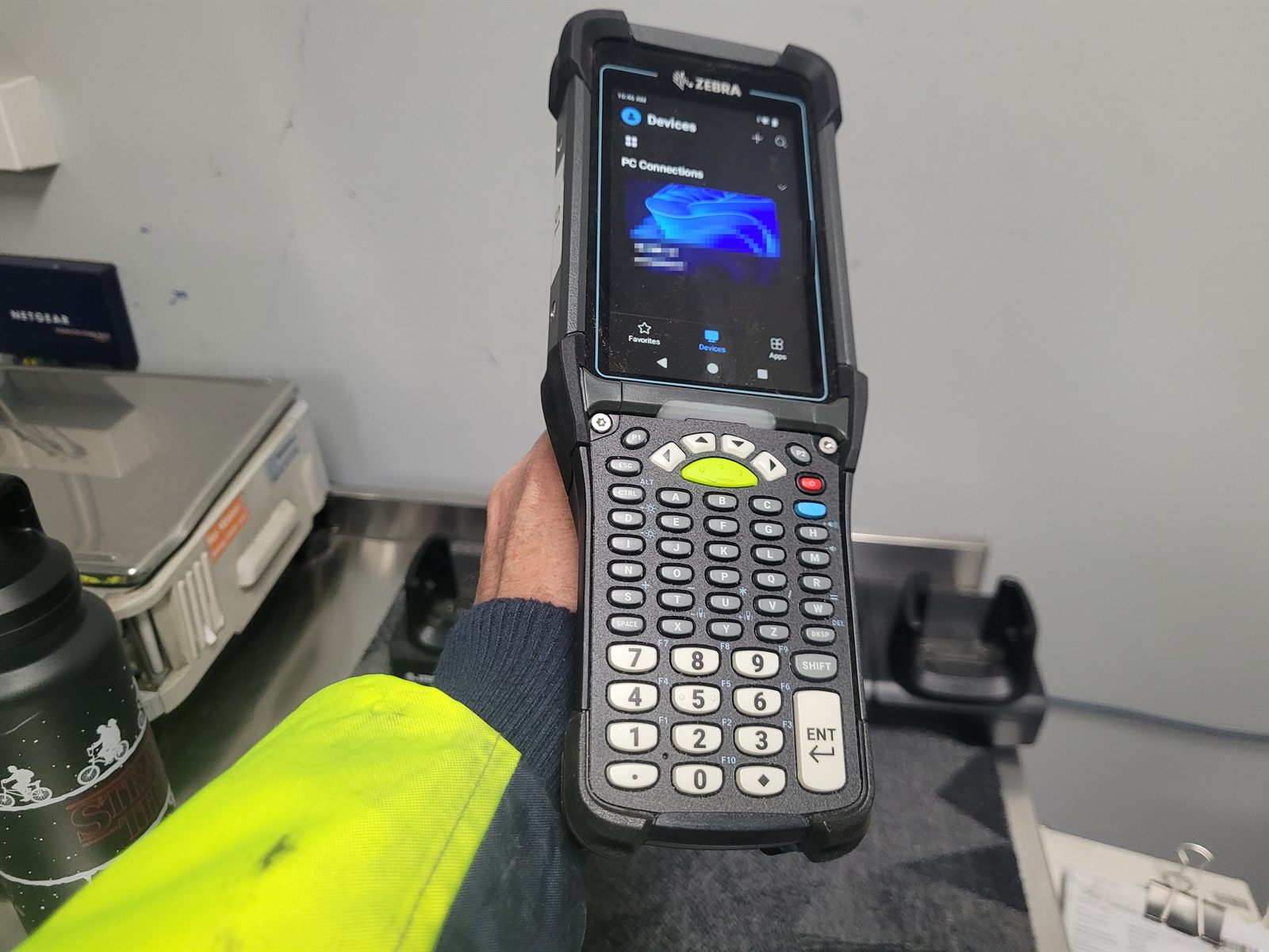 A Zebra MC9400 held in one hand with a hi-vis sleeve visible, showing the 53-key keypad and a connection screen with one line pixelated.