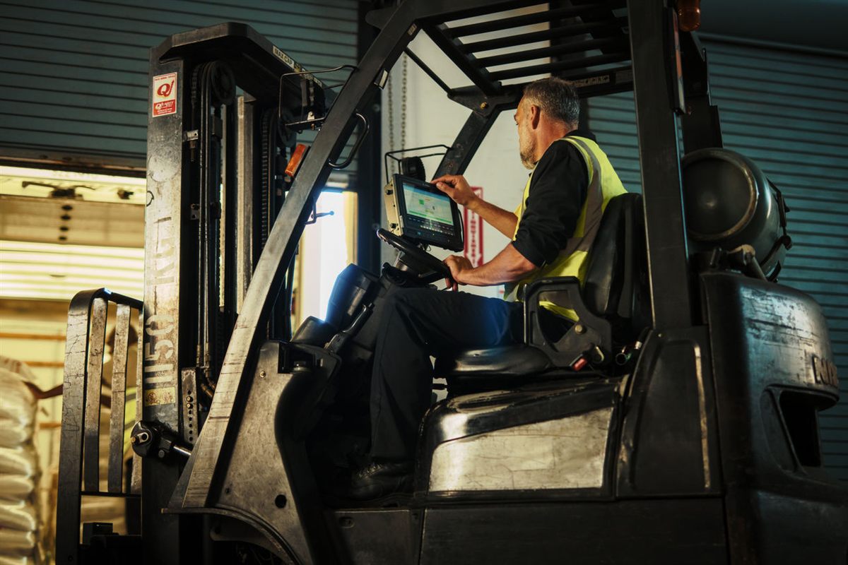 A forklift driver in a hi-vis vest, seated, one hand on a rugged touchscreen terminal bolted to the mast of the truck.
