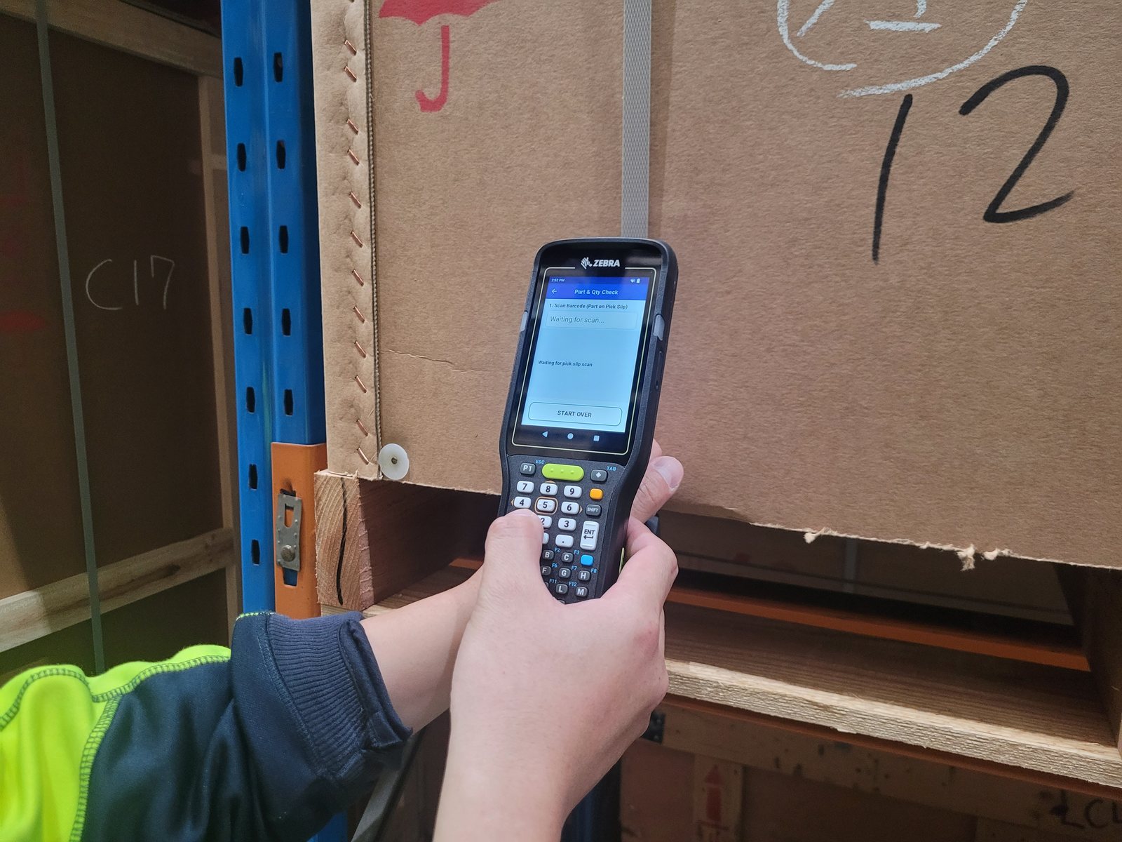 Two hands holding a Zebra MC3400 in a warehouse aisle in front of a strapped
                  timber crate, the screen waiting for the pick slip barcode to be scanned