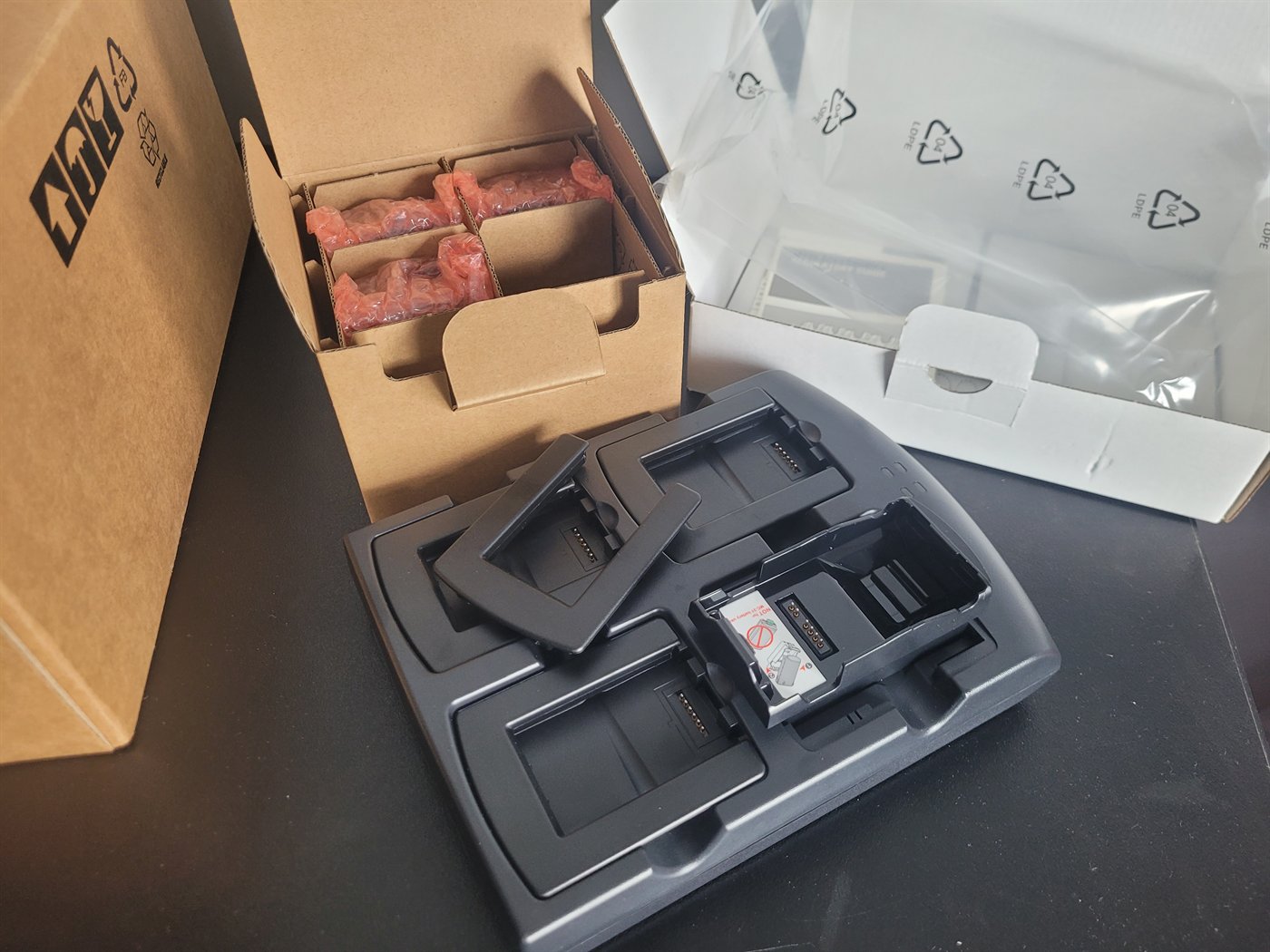 A Symbol four-slot battery charger opened out on a bench, with black plastic
                  adapter cups - one fitted into a bay and two loose beside it - and a small
                  carton of spare cups alongside.