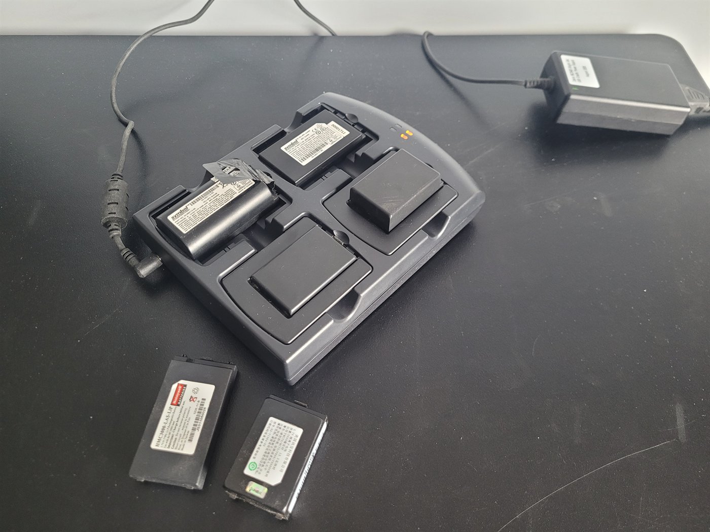 The same four-slot charger plugged in and running, with batteries of three
                  different shapes seated in its bays and two more on the bench beside it,
                  including a Honeywell-branded cell for an MC3000. Two amber charge lights
                  are lit.