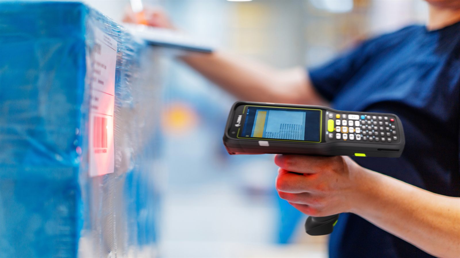 A warehouse worker holding a Zebra gun-grip mobile computer, its red aiming line
                  across a barcode label on a shrink-wrapped pallet, with a second worker checking
                  a clipboard further down the aisle.
