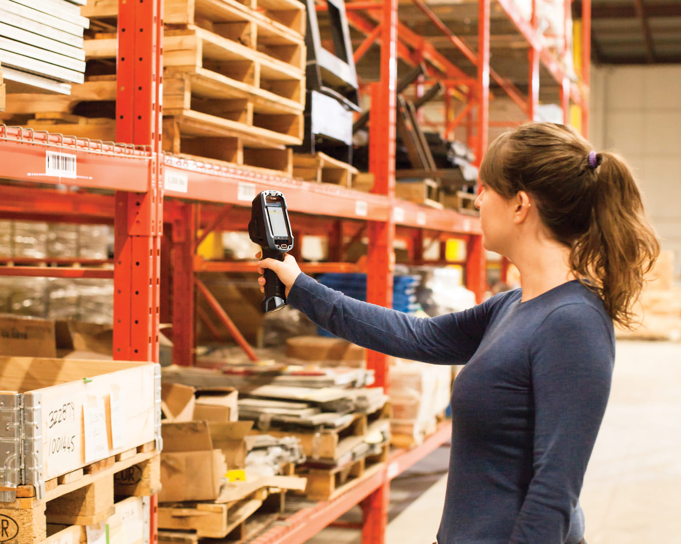 A warehouse worker reaching above head height to scan a barcoded label on the
                  beam of orange pallet racking, holding a rugged handheld scanner with a pistol
                  grip.
