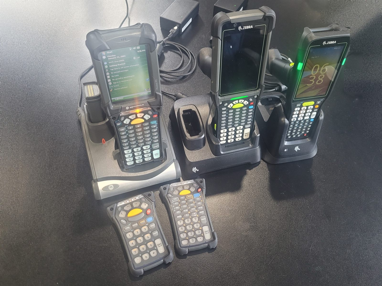 Three gun-grip mobile computers on a dark desk, each standing in its own
                    charging cradle. The left cradle has a spare battery in a well beside the
                    terminal, the middle cradle's well is empty, and two loose keypads lie in
                    front of them.