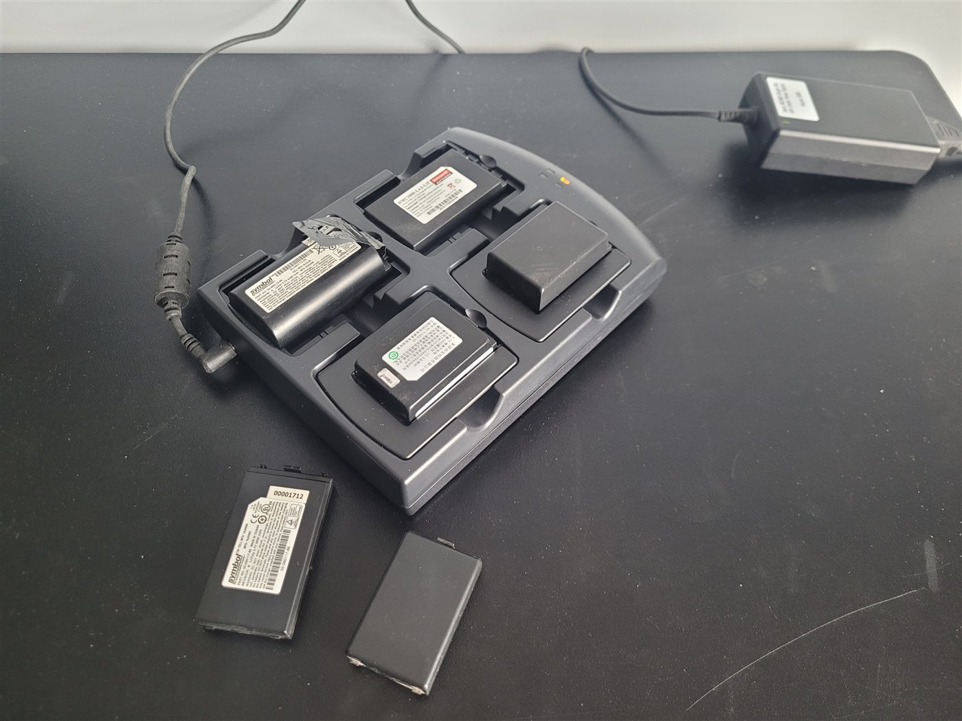 The four-slot charger running on a bench with four batteries of different sizes seated in its bays and two more beside it. The batteries are not included in the kit and are shown only to demonstrate what fits.
