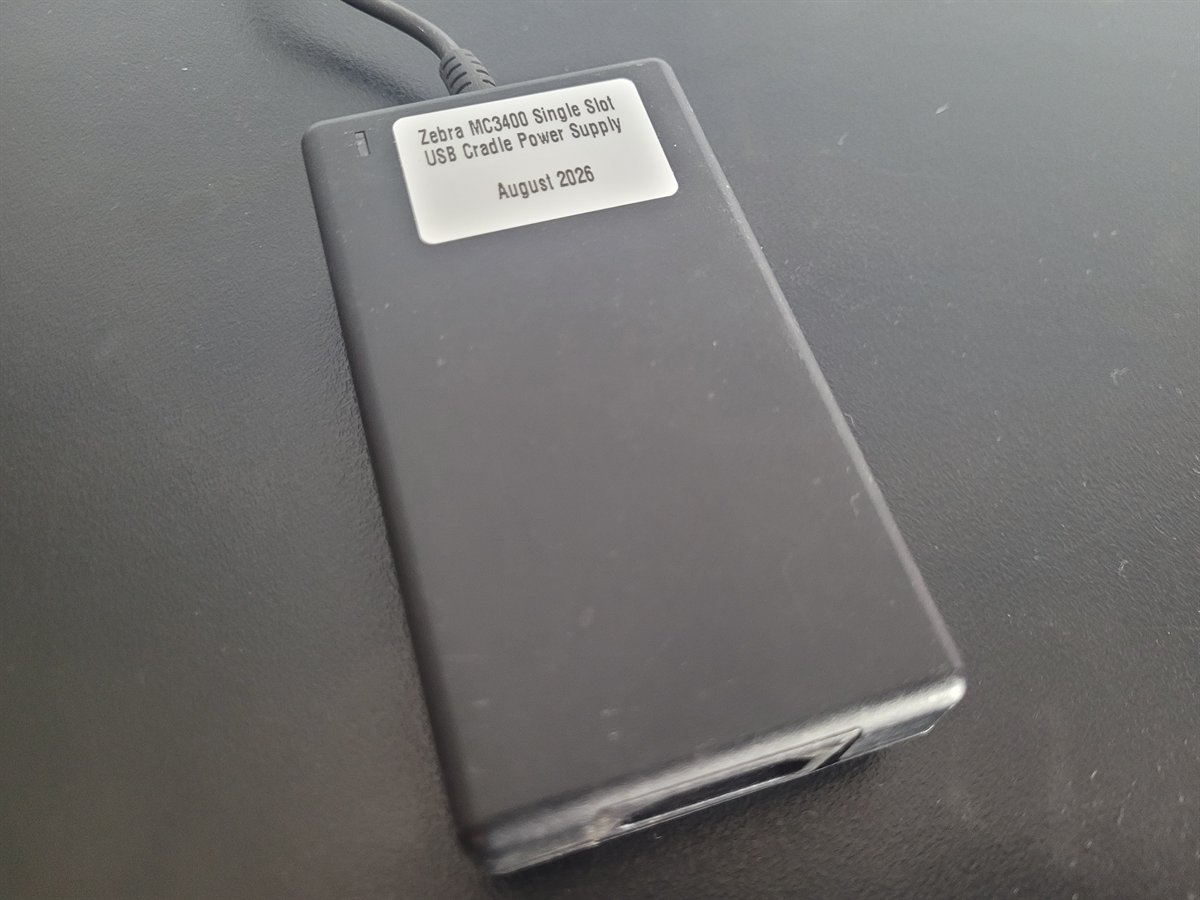 A black Zebra power supply brick on a bench, carrying a printed label reading
                  Zebra MC3400 Single Slot USB Cradle Power Supply, August 2026.